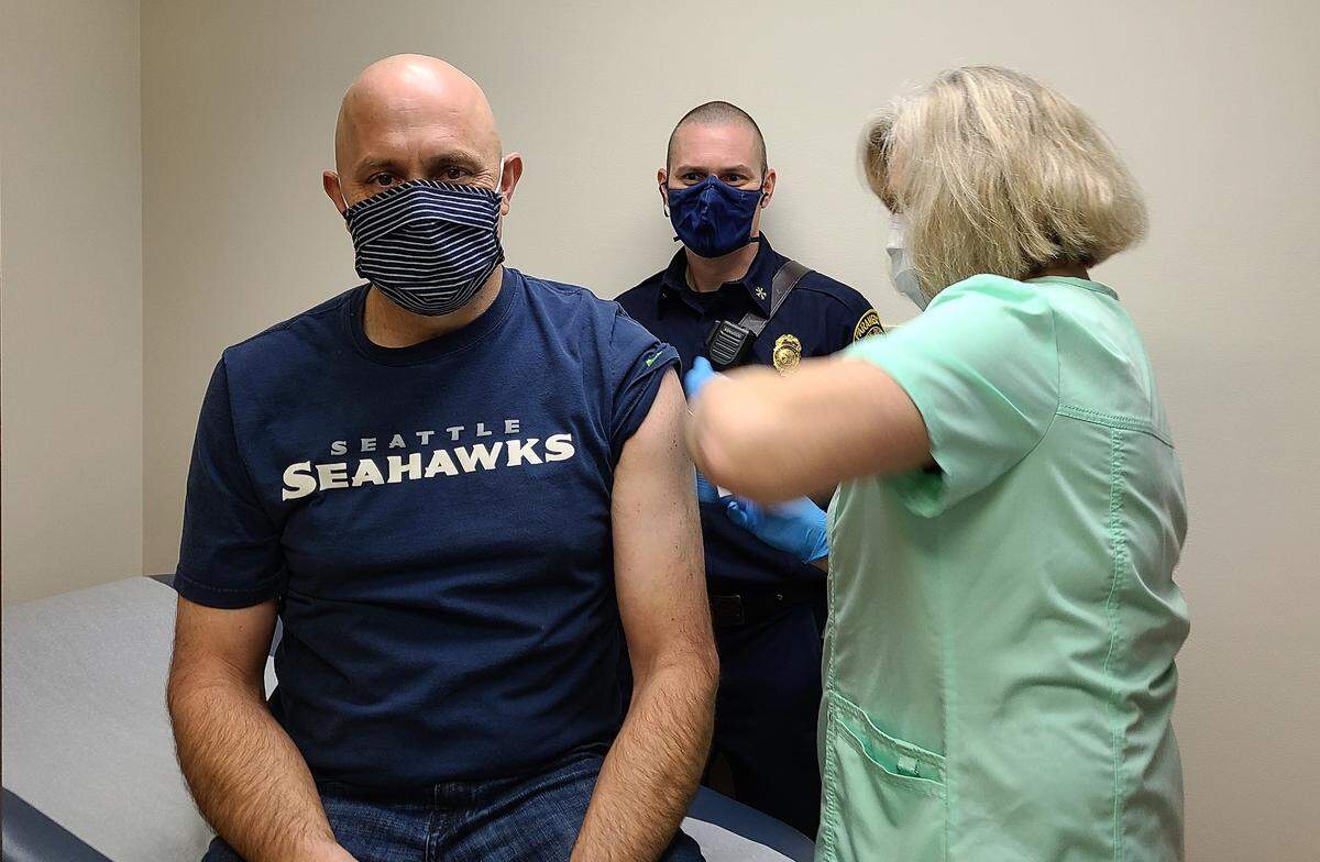 Kelly Devlin receives the first official vaccination at an EMS vaccine clinic Monday, Dec. 28, in Bellingham from Nancy VanderPol, certified medical assistant with PeaceHealth Medical Group Pediatrics, as Micah Quintrall waits to be the second in line. Devlin and Quintrall are paramedics/firefighters with the Bellingham Fire Department.