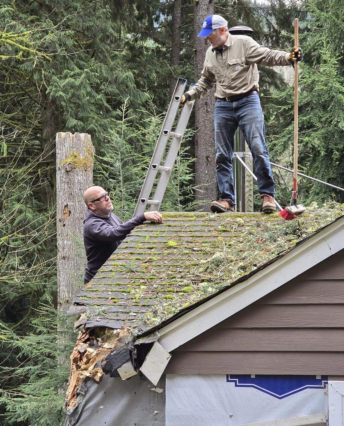 Sudden Valley homeowner Dwayne Campbell, left, and neighbor Sam Shahan, right, help clear tree limbs and other storm debris off Campbell’s roof on Saturday, Nov. 23, in the forested community east of Bellingham. A 140-year-old hemlock clipped the corner of Campbell’s home as it fell on the night of Nov. 19 during a bomb cyclone windstorm.