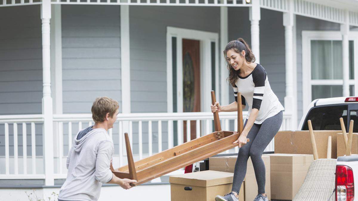 A young couple moving into a house or apartment. Their pickup truck is full of cardboard boxes and furniture. They are lifting a small table.