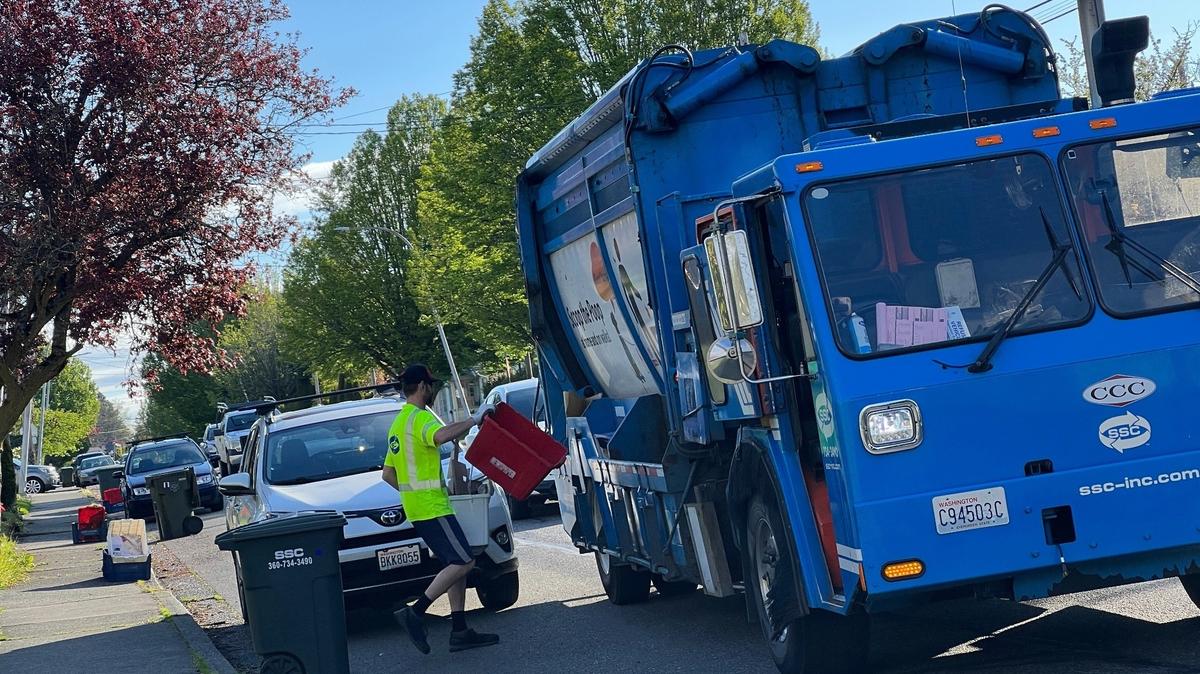 A worker for Sanitary Service Co. empties recycling bins into a truck on F Street near Halleck Street on Tuesday, May 9, 2023, in Bellingham, Wash. New single-bin recycling containers can be loaded onto trucks with mechanical arms, and reduce injuries among drivers who currently must leave the truck to lift and empty the bins themselves.