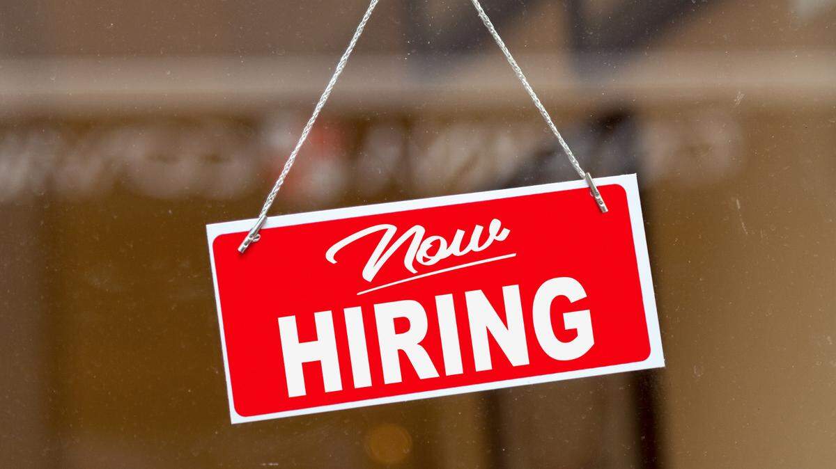 Red sign hanging at the glass door of a shop saying: “Now hiring”.