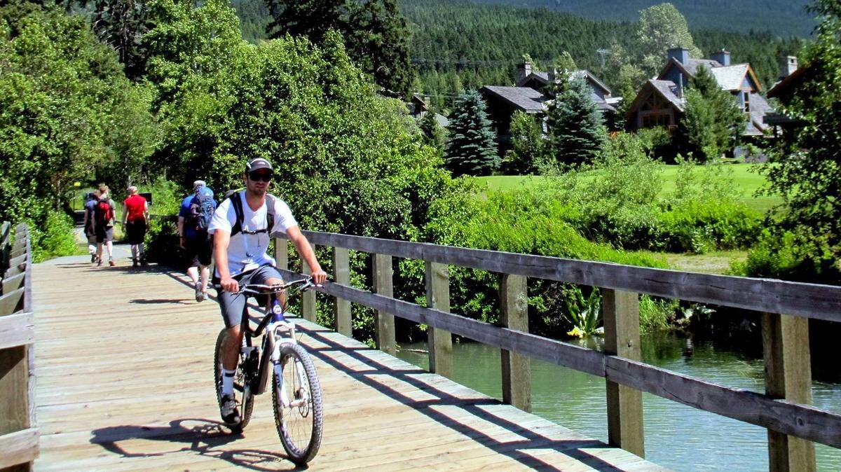 A bicyclist and walkers travel a short boardwalk portion of the 24-mile Valley Trail in Whistler, British Columbia. The paved trail winds through woods and parks, past small lakes and homes.