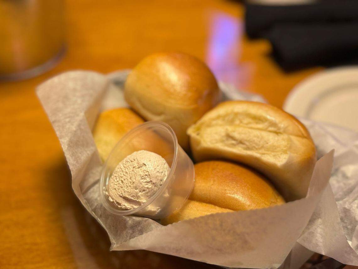 A close-up of a basket of golden-brown Texas Roadhouse rolls lined with wax paper. A small container of cinnamon butter is nestled among the rolls.