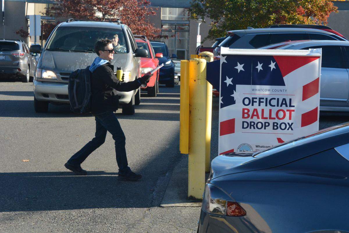 A voter on foot places their ballot in a drive-up collection box as drivers wait outside the Whatcom County Courthouse.
