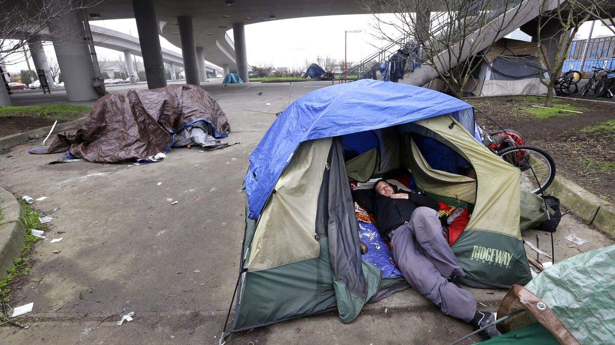 FILE - In this Feb. 9, 2016 file photo, a man lies in a tent with others camped nearby, under and near an overpass in Seattle. Microsoft pledged $500 million to address homelessness and develop affordable housing in response to the Seattle region’s widening affordability gap. The company, which plans a news conference Thursday, Jan. 17, 2019, will split the funds three ways. (AP Photo/Elaine Thompson, File)