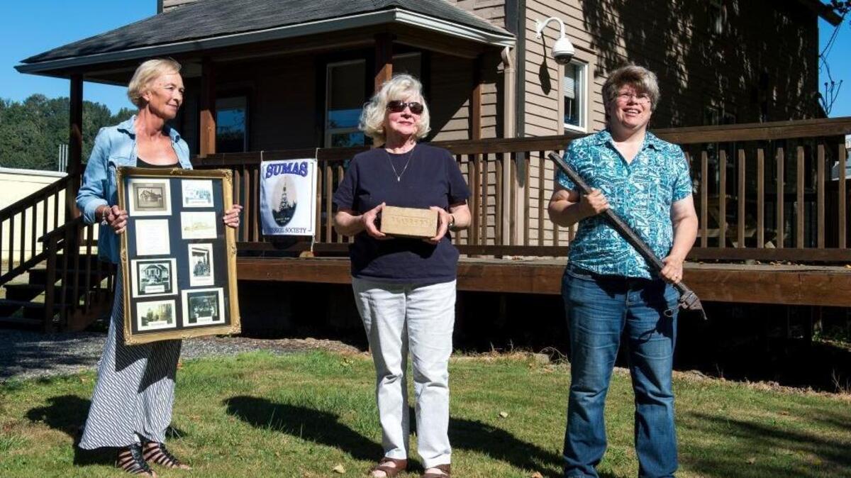 From left to right, Sumas Historical Society members Lizette Custer, board member, Gail Kihn, treasurer, and Deborah Morgan, president, hold artifacts in front of the future museum of the Sumas Historical Society on Thursday, Aug. 18. Custer is holding a collection of photos of the historical John C. Gillies home where she currently lives. Kihn is holding a Clayburn brick, made in nearby Clayburn, B.C., which was used in the construction of many buildings in Sumas. Morgan is holding a rifle she found in 1978 with a metal detector at the site of one of Sumas' original houses.