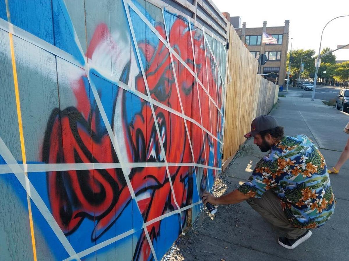 Shawn Cass uses tape to perfect the lines in the checkerboard background of the mural he is painting on Schweinhaus Biergarten’s fence.