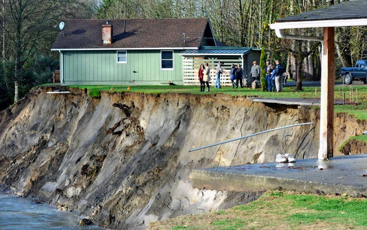 Local residents and government officials stand next to Michael Taxdahl's house in Lyman on Monday, one of several which is threatened by erosion following the Skagit River flood on Thanksgiving. In the foreground, the concrete porch of another home is undermined by the failing embankment.