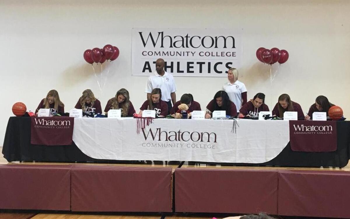 Mount Baker’s Kylee Engholm, from left, Lynden Christian’s Shyann Brandsma and Emma Stump, Blaine’s Kjirsten Larson, Cornerstone Christian’s Elissa Vreugdenhil, Meridian’s Kiana Gray, Lynden’s Mariah Gonzalez, Nooksack Valley’s Rachel Sande and Sehome’s Samantha Hruby sign letters of intent to play basketball for Whatcom Community College on Saturday at Orca Pavilion, as coach Kevin Davis and assistant Courtney Shilling watch.